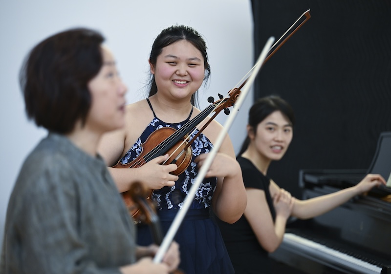 A violinist smiles while holding her violin to her chest with her accompanist behind her, while listening to a professor in a master class