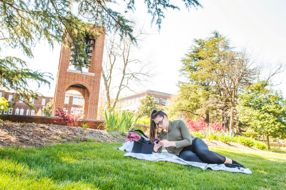 While using her phone, a young woman with a long ponytail and glasses sits on a blanket on the grass while the UNCG campus buildings and trees surround her.